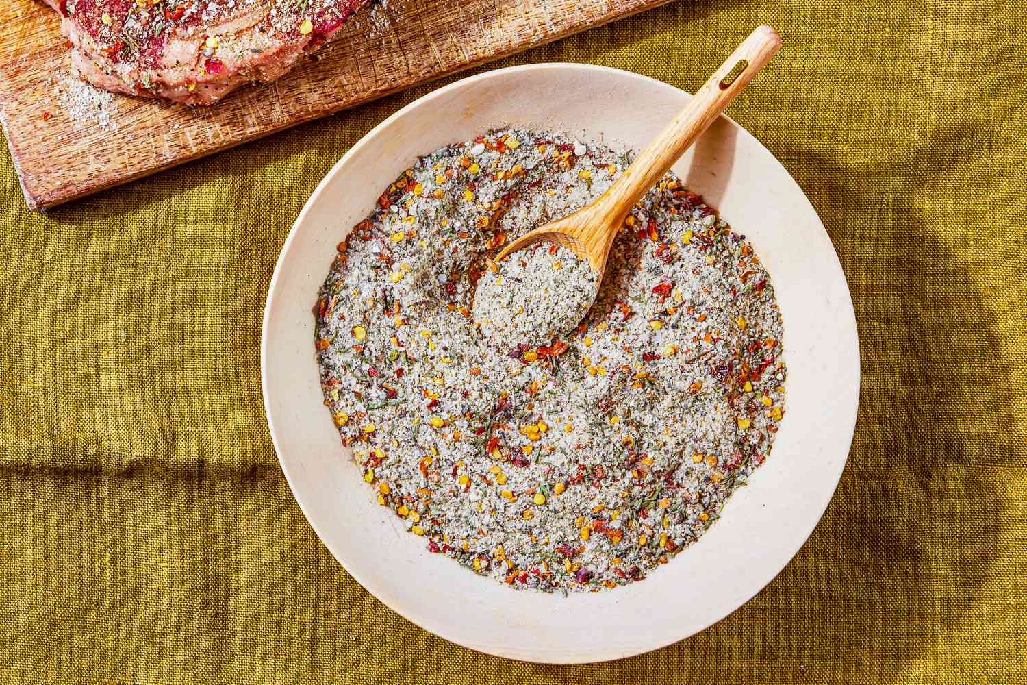 Bowl of Steak Seasoning With a Spoon Next to Cutting Board With a Seasoned Raw Steak