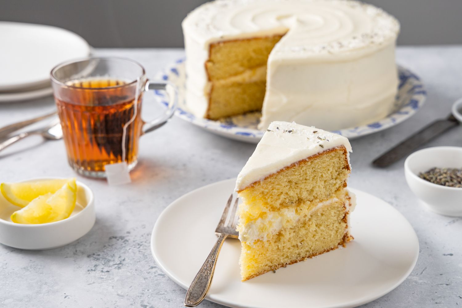 Slice of Lemon Lavender Cake on a Plate with a Fork Surrounded by a Dish of Lemon Wedges, a Small Bowl of Lavender, Cup of Tea, and Leftover Cake in Background