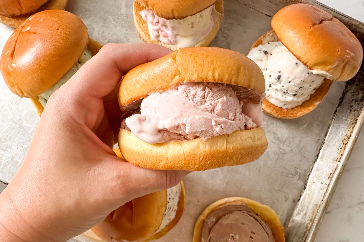 Hand holding a brioche sandwich with ice cream, with similar sandwiches on a tray in the background