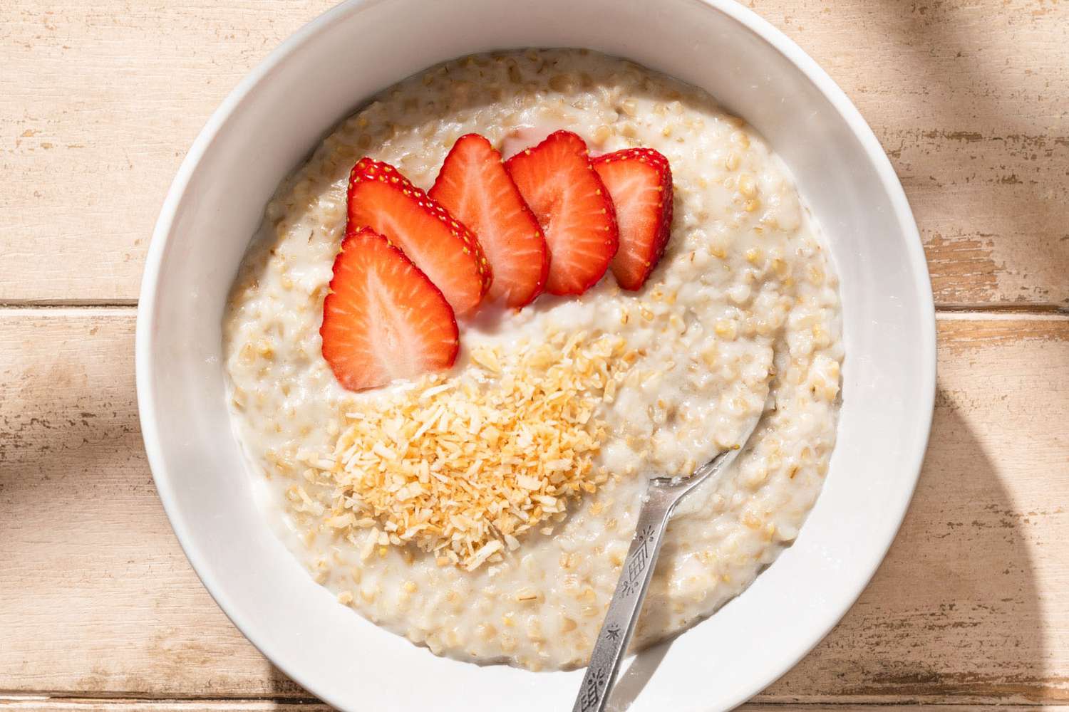 Coconut oatmeal topped with sliced strawberries and toasted coconut, served in a bowl with a spoon.