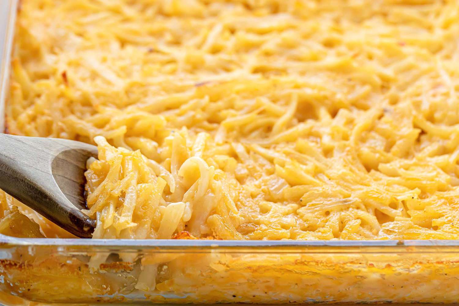 A side view of a glass casserole dish of potato casserole without canned cream soup. A wooden spoon is partially in view and scooping a serving out of the casserole dish.