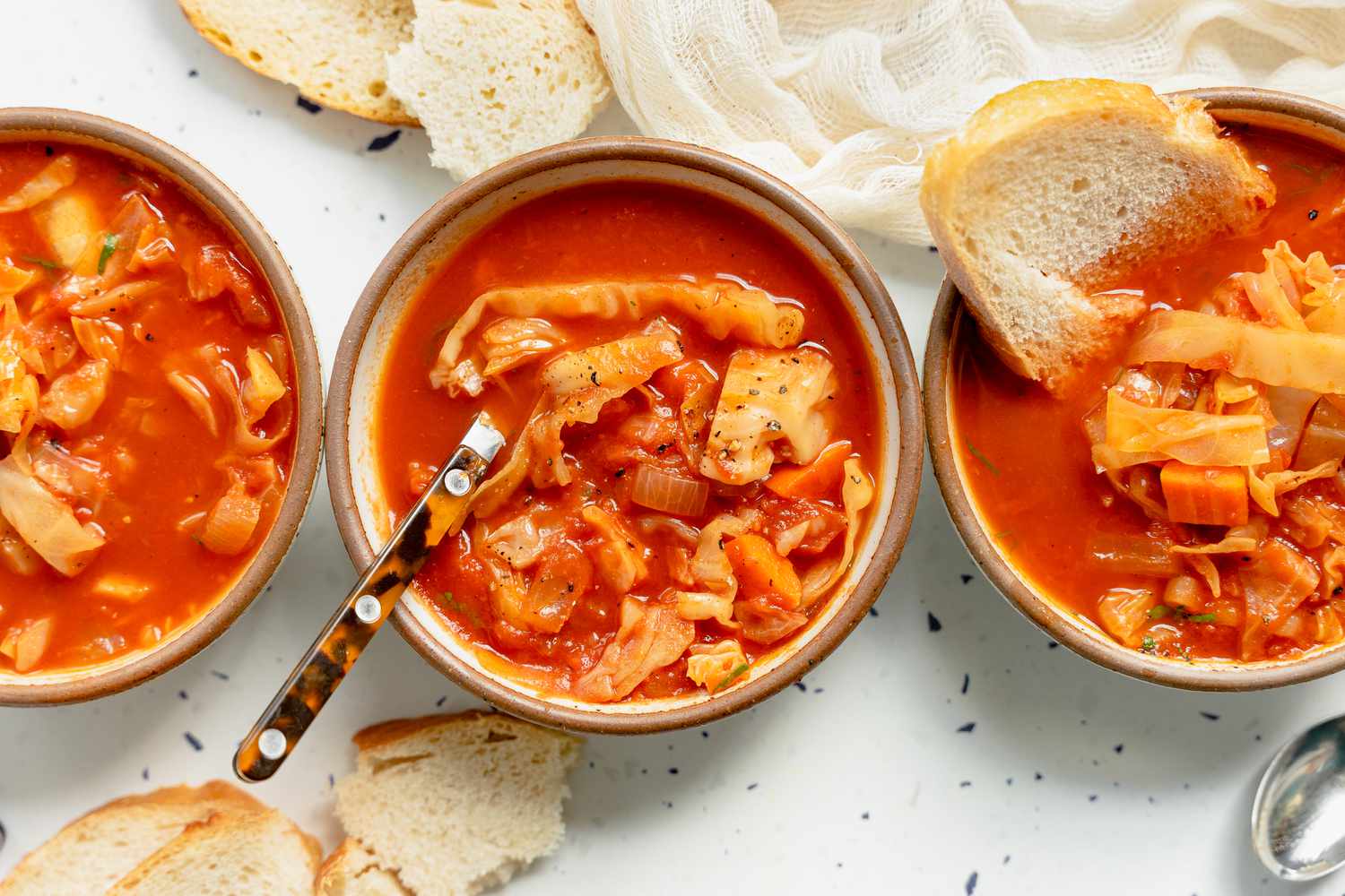 Three Bowls of Sweet and Sour Cabbage Soup. One Bowl Has a Spoon, Another Has a Slice of Bread. Bowls Are Surrounded by Slices of Bread and a Kitchen Linen