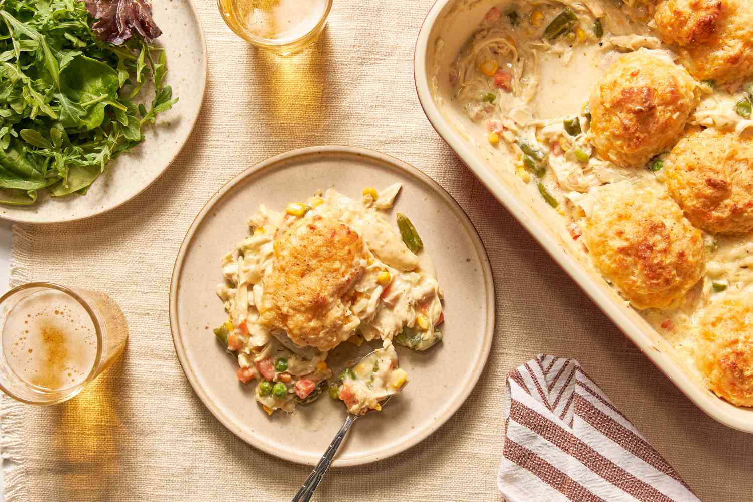 Serving of Chicken Cobbler With Cheddar Bay Biscuit Top on a plate at a table setting with a plate of salad greens, glasses of cider, and a casserole dish with cobbler