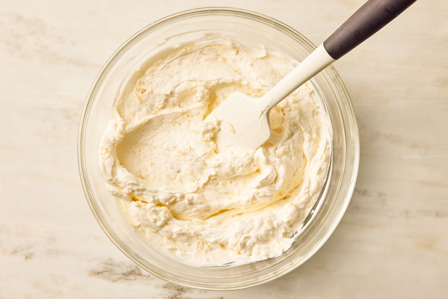 Overhead view of a glass mixing bowl with the ingredients of the frosting being mixed with a spatula for Pig Pickin' Cake recipe