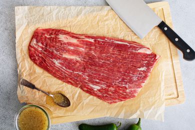 Flank Steak on butcher paper on a cutting board with a butcher knife, marinade in a small bowl on the side with a spoon