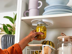 Hand Holding a Closed Jar up to a Jar Opener Installed under One of the Cabinet Shelfs. In the Surroundings, a Cabinet Setting With Stacks of Plates, a Salad Bowl, Mugs, a Container of Flour, and a Basket of Fruit and a Potted Plant Next to the Cabinet. 
