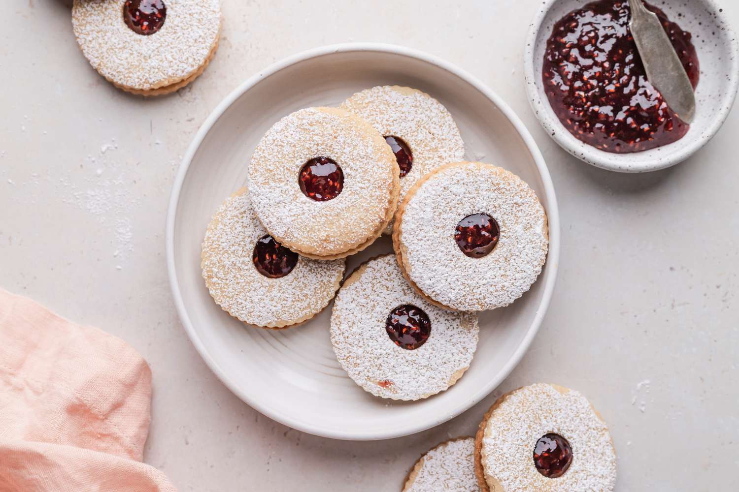 Jam filled sandwich cookies on a plate with additional cookies and small saucer of jam set around the plate.
