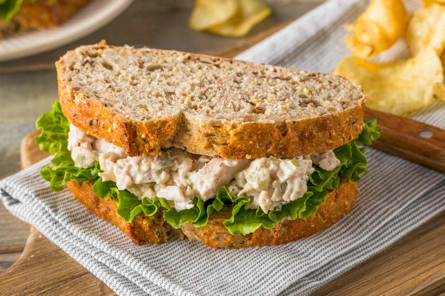 Chicken salad sandwich on whole-grain bread, with lettuce and potato chips in the background.