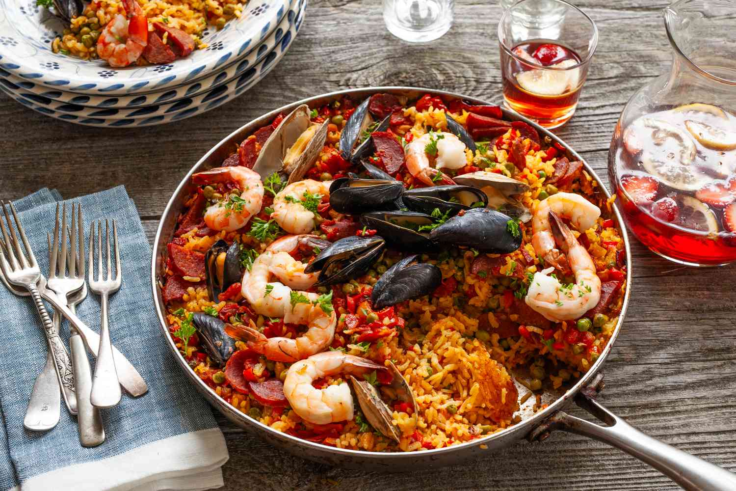 A pan of seafood paella on a wooden table with utensils, plates, and a glass of sangria nearby