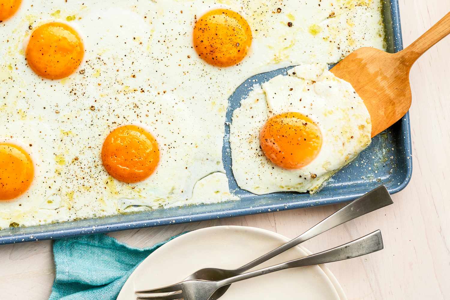 Wooden Spatula Lifting a Sheet Pan Fried Egg From a Baking Pan, and Next to the Sheet Pan, a Stack of Plate With Utensils and a Kitchen Towel