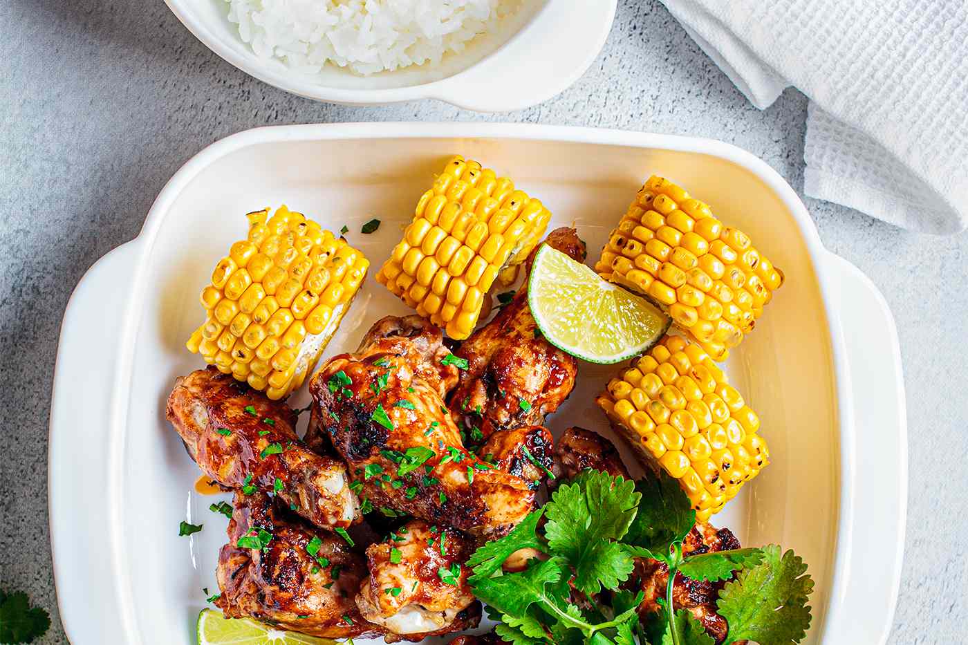 A plate with corn on the cob and grilled chicken wings glazed with a reddish sauce. Cilantro and lime wedges garnish the food.