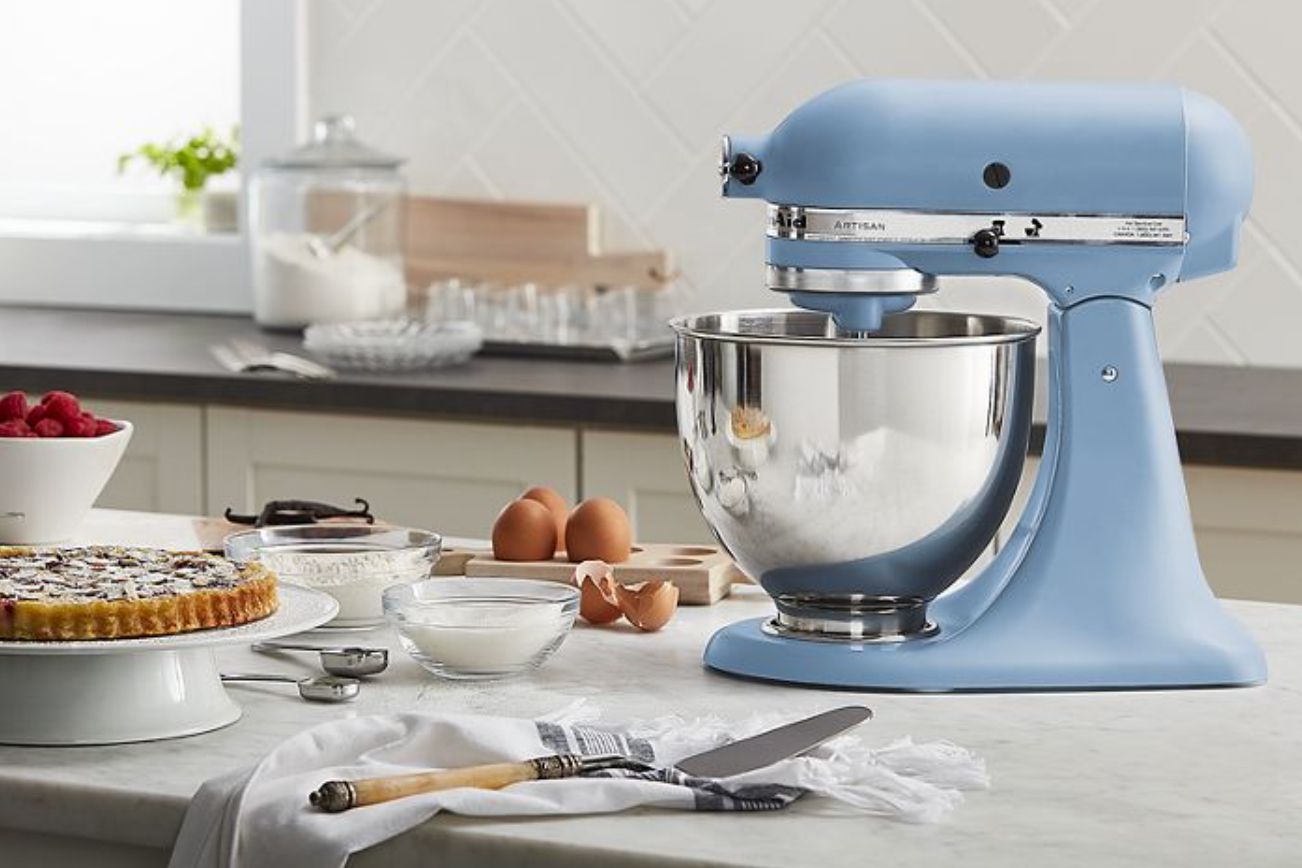 Side view of a light blue stand mixer in the lowered position along with it's metal bowl. Shown on a kitchen counter along with various ingredients, baking tools and a tart on a cake stand