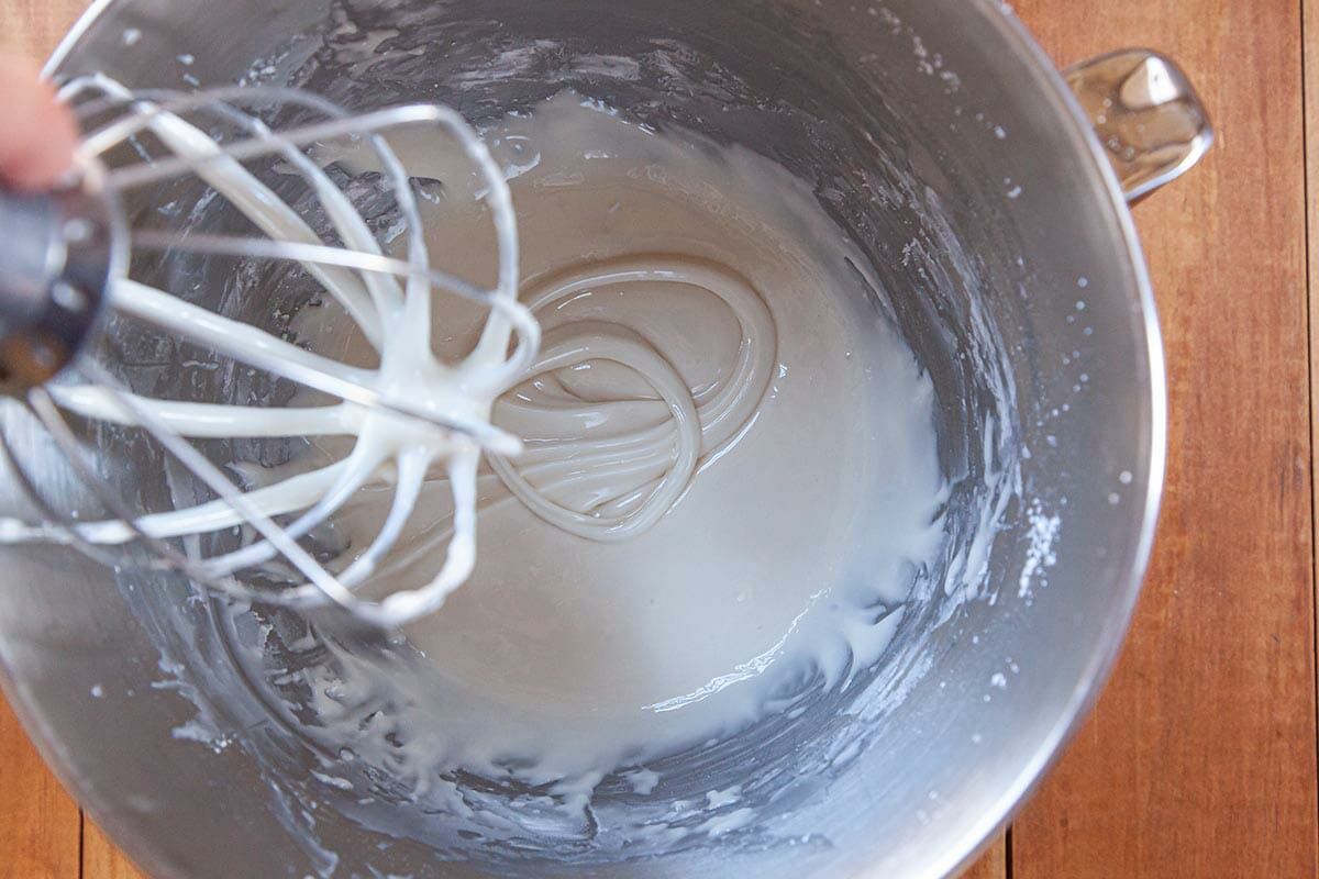 A whisk is lifted out of a stand mixer filled with Royal icing for sugar cookies.