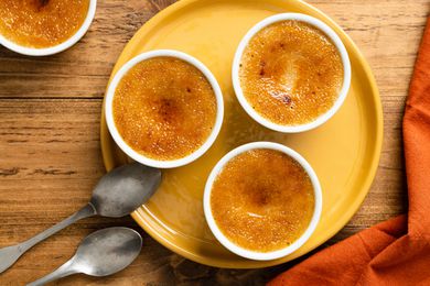 Overhead Shot: Multiple Ramekins Filled with 3-Ingredient Pumpkin Spice Crème Brûlée Sitting on a Plate Next to Two Spoons, a Table Napkin, and Another Ramekin with More Pumpkin Spice Creme Brulee