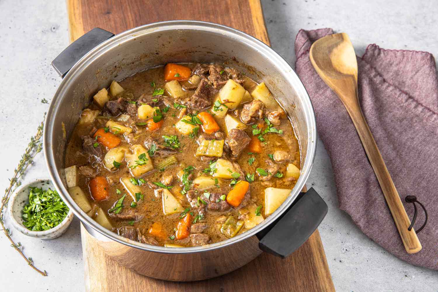 Pot of Easy Beef Stew on a Long Wooden Board, and Next to It, a Bowl of Parsley and a Sprig of Thyme on the Left and a Kitchen Towel and a Wooden Spatula on the Right 