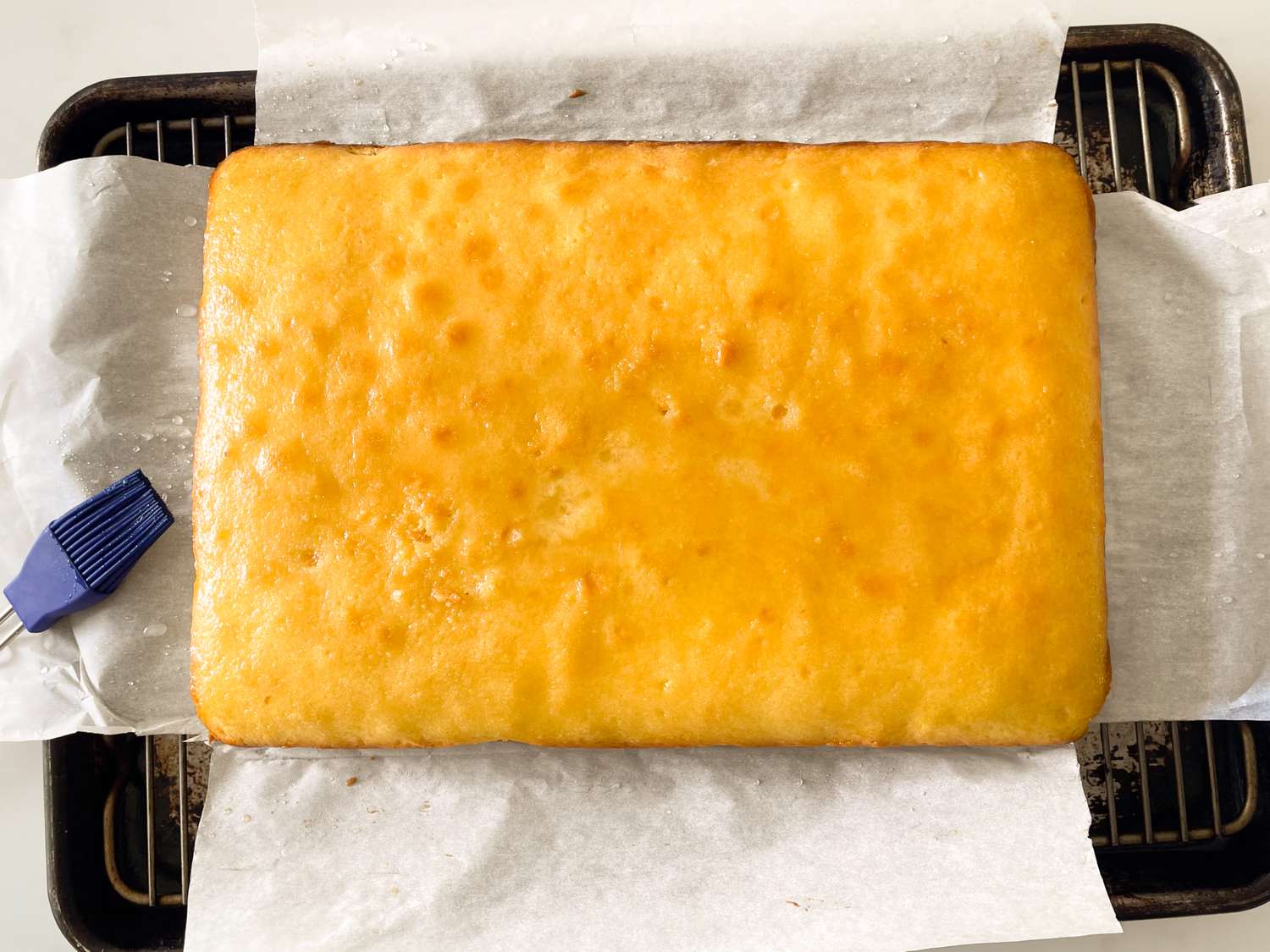 A baked rectangular cake cooling on parchment paper - Mary Berry’s Lemon Drizzle Traybake Cake