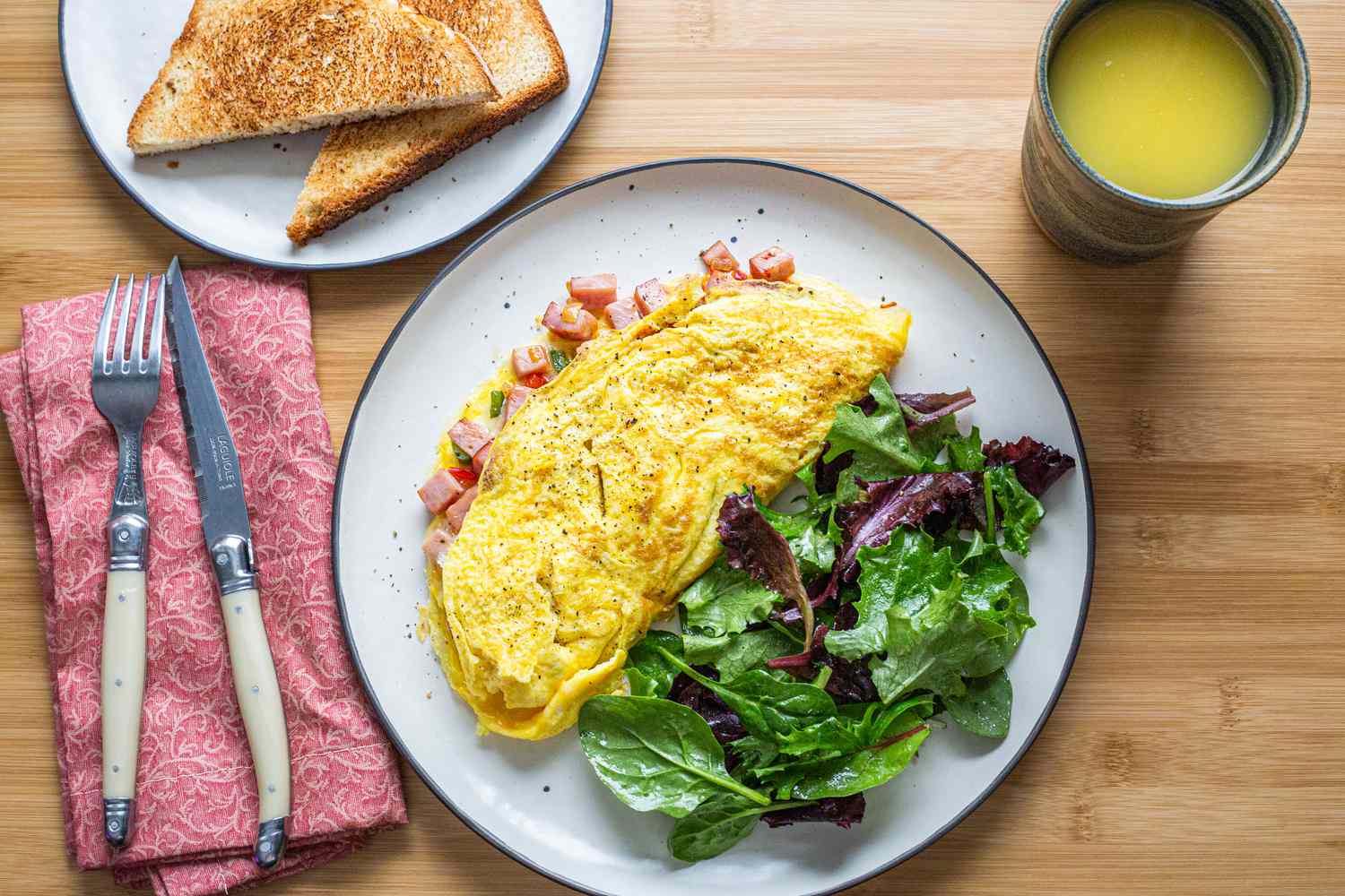 Denver Omelet with Mixed Greens on Plate Next to Cup of Juice, Plate of Toast, and Utensils on Napkin