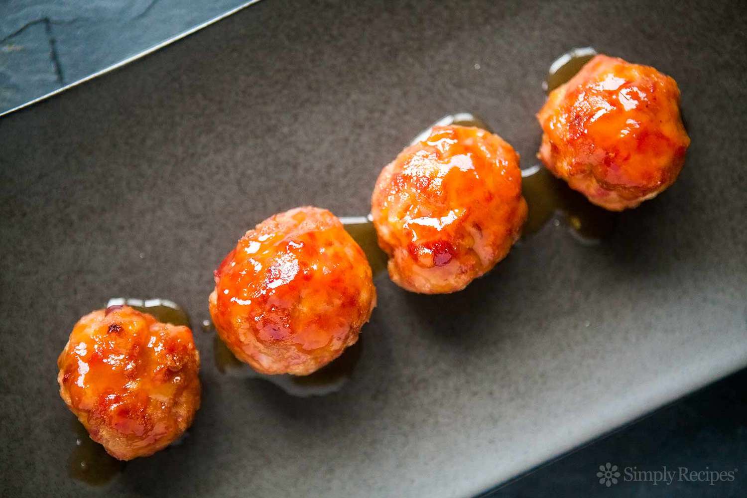 Four glazed meatballs arranged in a line on a dark plate resting on a drizzle of sauce
