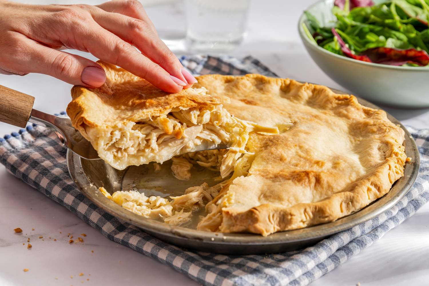 A person serving a slice of chicken pie from the whole pie with a salad bowl in the background