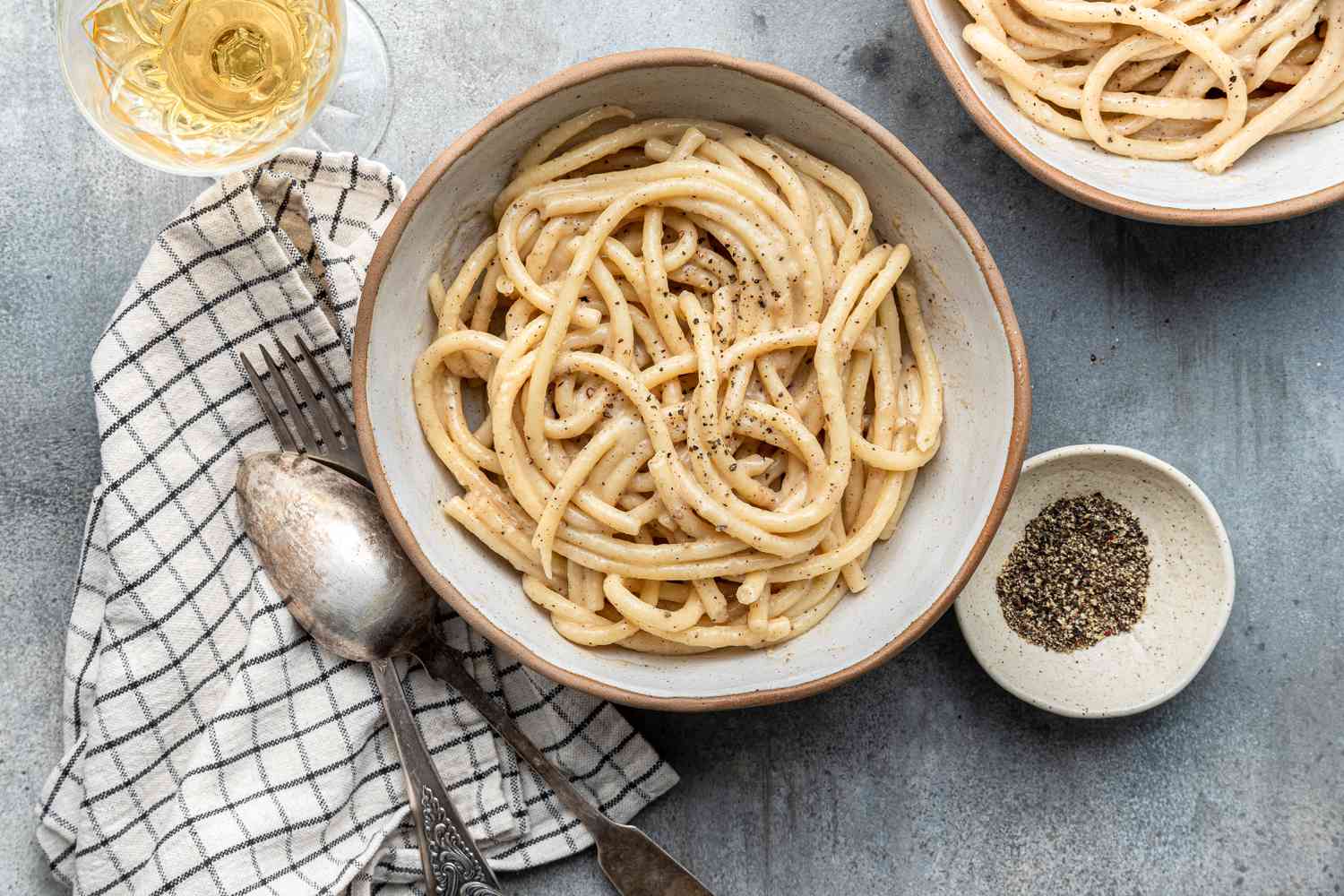 Bowl of Brown Butter Cacio e Pepe Next to a Fork and Spoon on a Kitchen Towel, a Glass of Wine, a Bowl with More Cacio e Pepper, and a Small Bowls with Cracked Black Pepper 