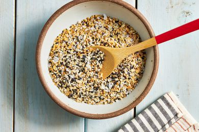 Bowl of Everything Bagel Seasoning With a Wooden Spoon and Next to a Black and White Stripped Kitchen Towel