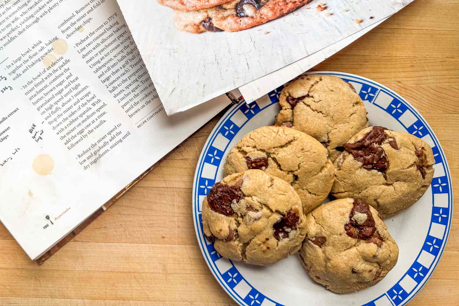Plate of chocolate chip cookies next to an open cookbook on a wooden surface