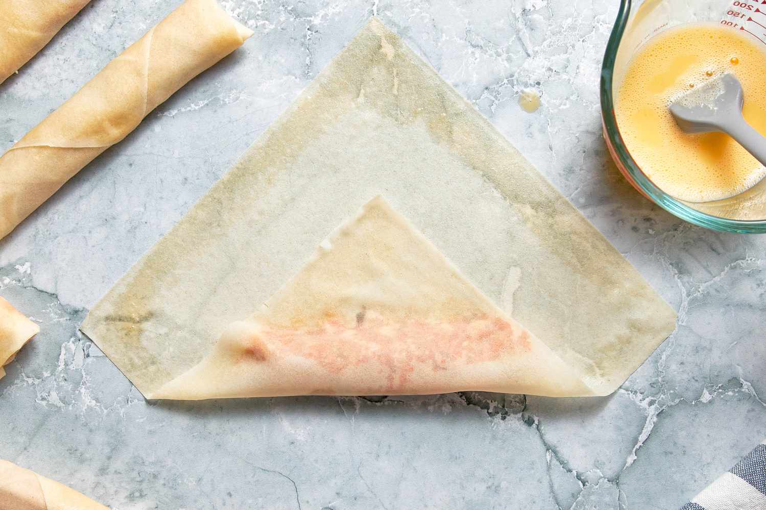 Bottom of Egg Roll Wrapper Folded Over the Filling, and in the Surroundings, and in the Surroundings, Rolled Lumpia, a Pyrex Measuring Cup With the Egg Wash, and a Kitchen Towel