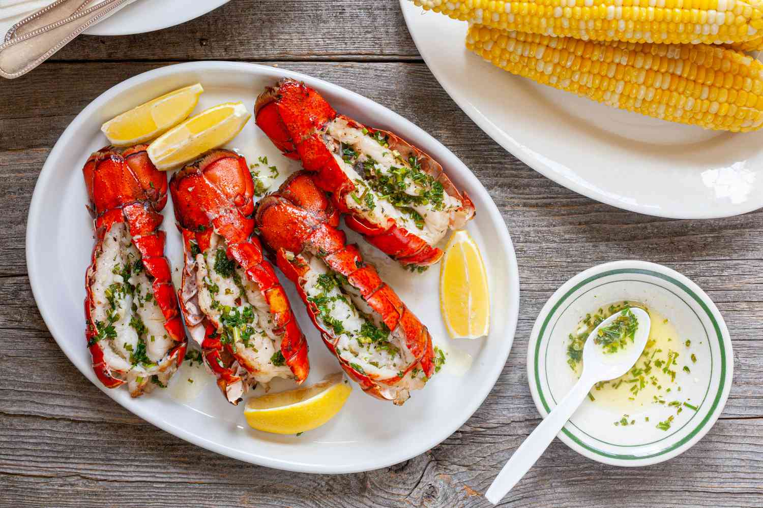 Platter of Grilled Lobster Tails with Lemon Wedges Surrounded by a Bowl of Garlic Herb Butter and a Plate of Corn 