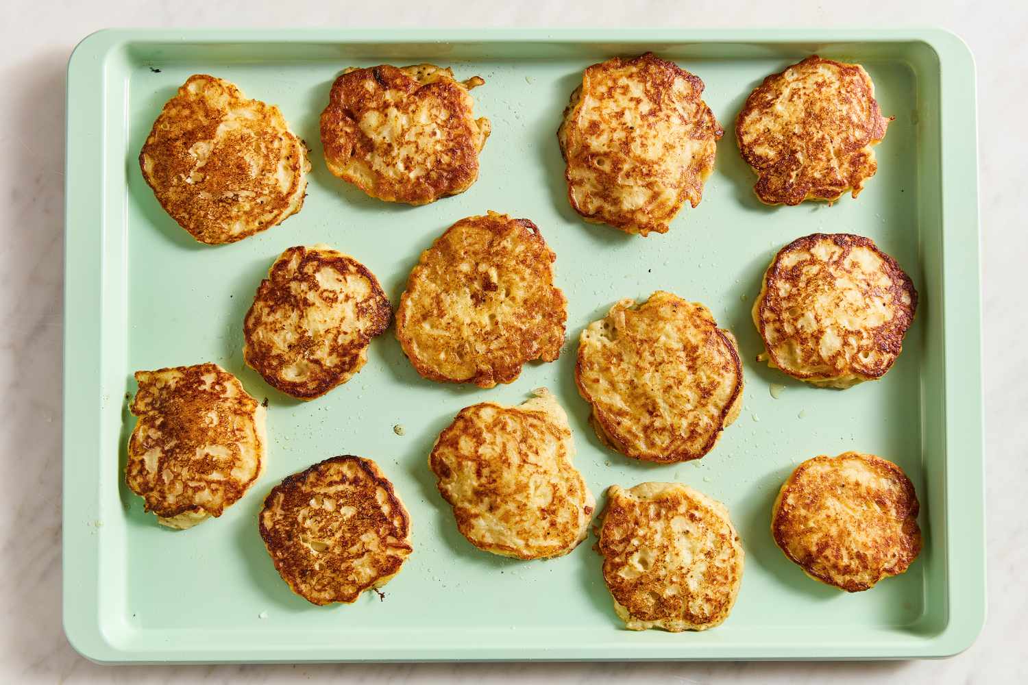 Overhead view of a light green baking sheet with thirteen cooked potato pancakes to keep warm in oven from Boxty (Irish Potato Pancakes) recipe