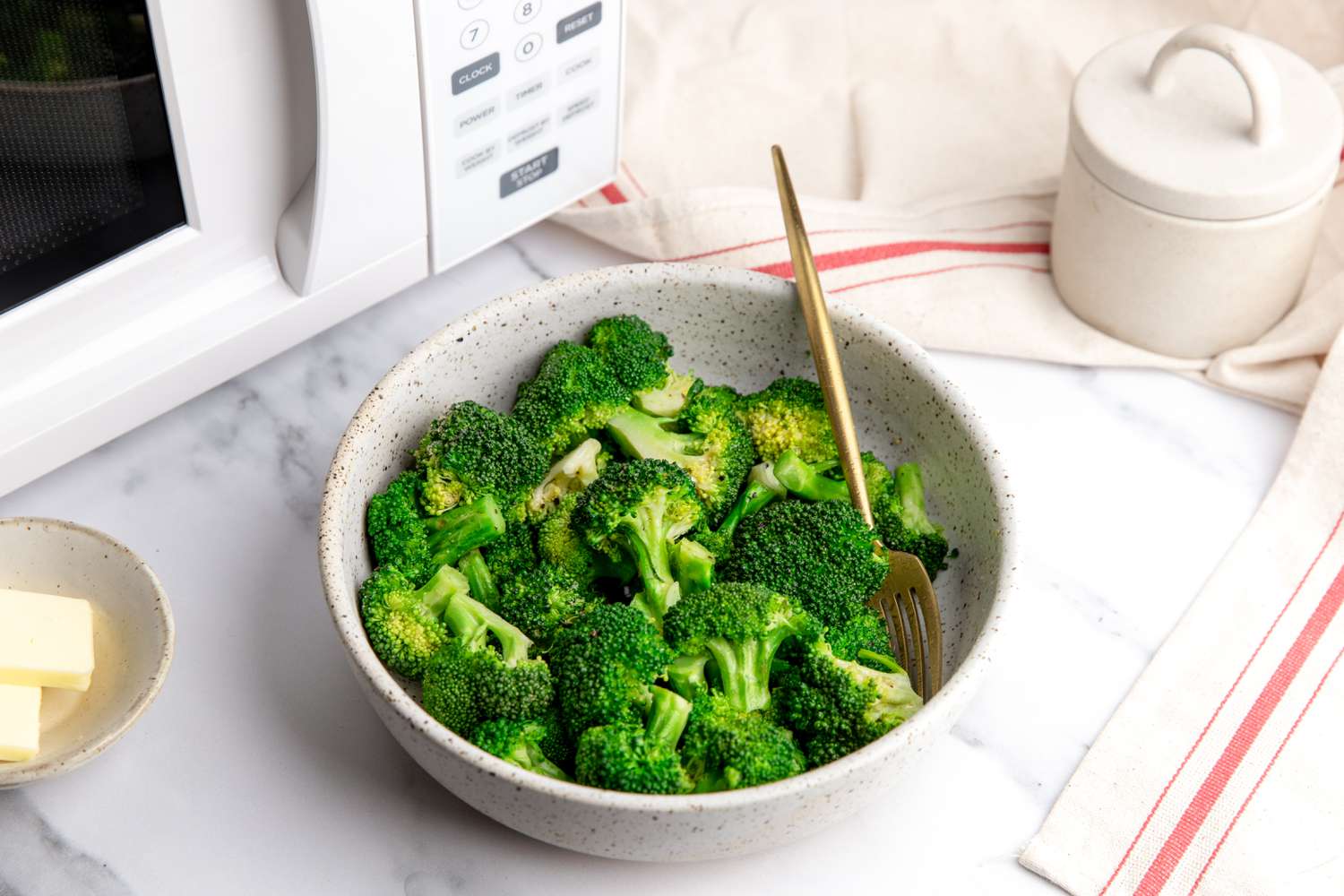 Bowl of Microwave Steamed Broccoli with a Fork, and in the Background, a Counter Setting with a Microwave, a Container of Salt, a Small Saucer with Slices of Butter, and a Kitchen Towel for How to Steam Broccoli in the Microwave