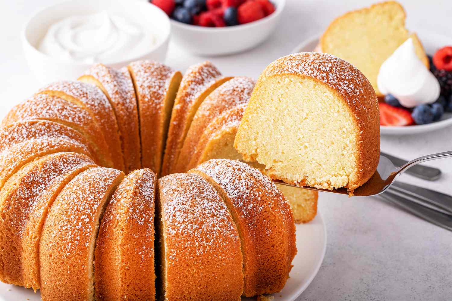Bundt cake with a slice being served accompanied by bowls of whipped cream and fruit