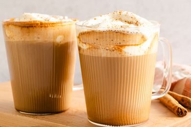 Two mugs filled with latte topped with whipped cream sprinkled with spices and placed on a wooden surface
