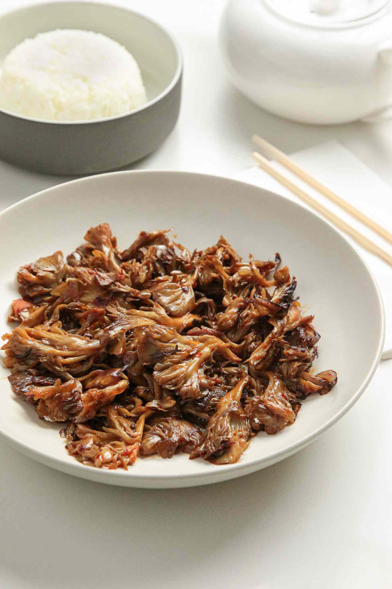 Stir-Fried Maitake Mushrooms with Garlic and Chile Oil in a Plate Next to a Pair of Chopsticks, a Bowl of Rice, and a Tea Kettle