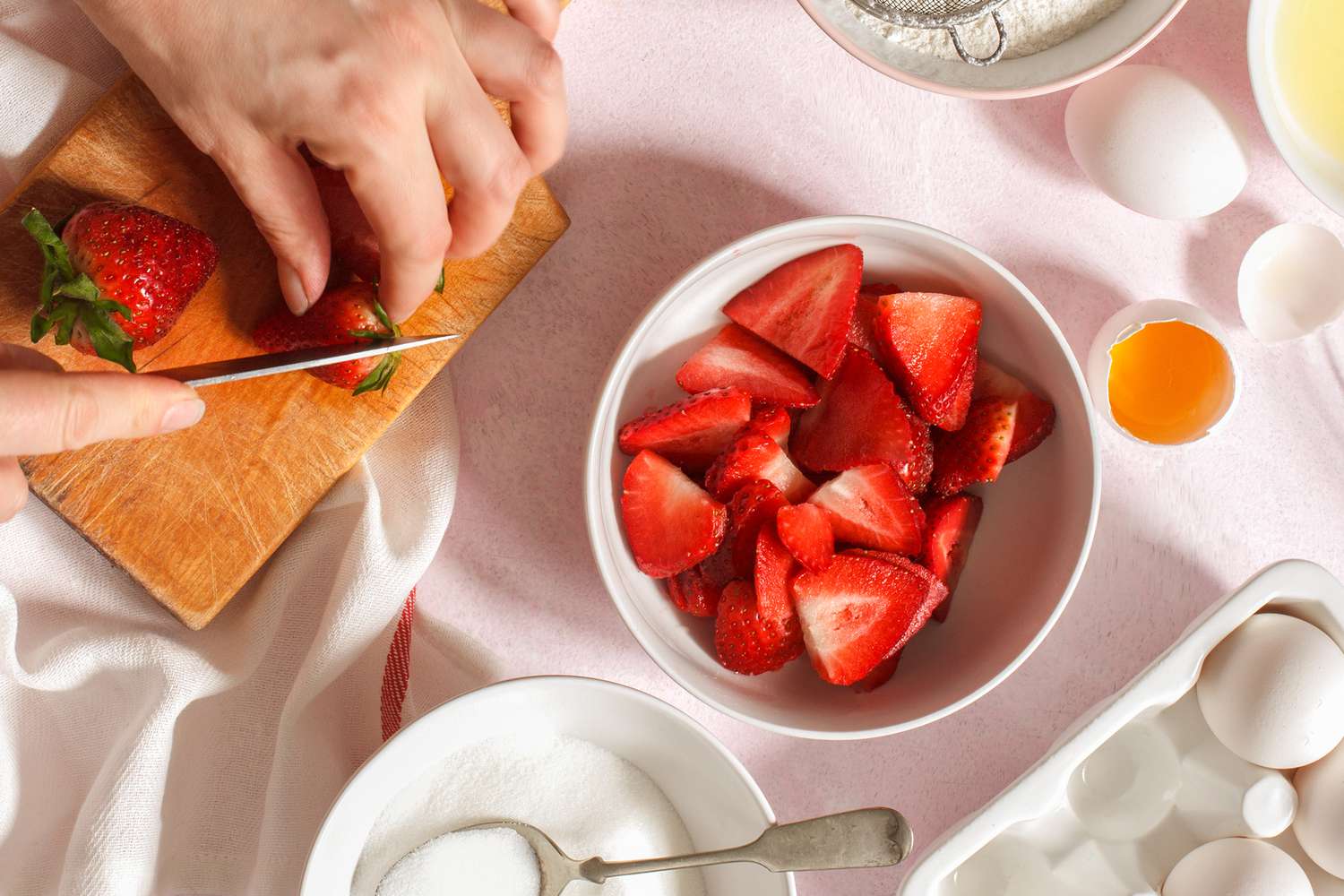 Slicing strawberries on a wooden board next to a bowl of strawberries, surrounded by baking ingredients