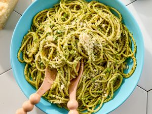 A bowl of pasta coated with pesto garnished with grated cheese with wooden utensils placed on top