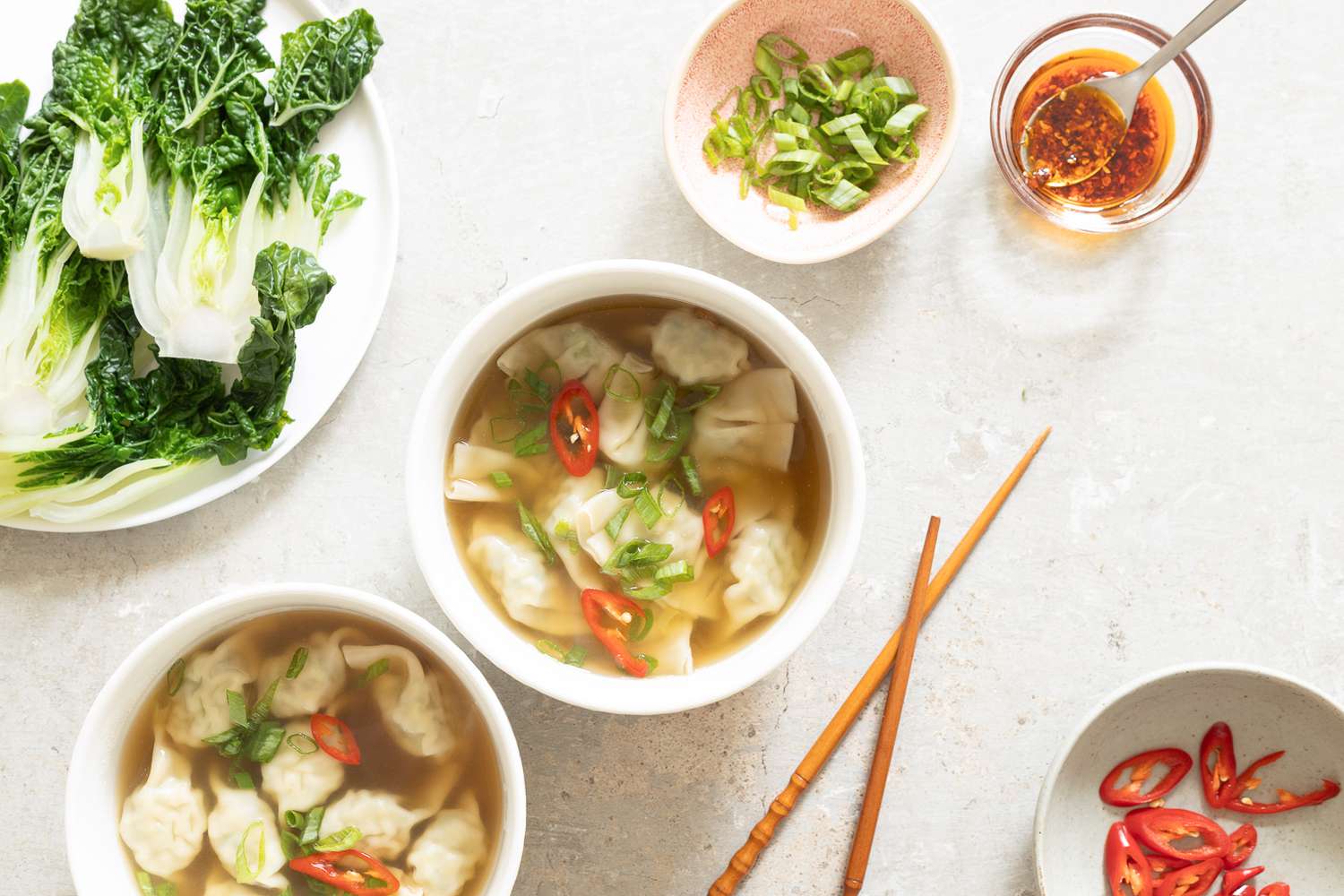 wanton soup in two bowls surrounded by toppings (a plate of bok choy, a small bowl of scallions, a small bowl of chili crisp, and a bowl with sliced peppers)