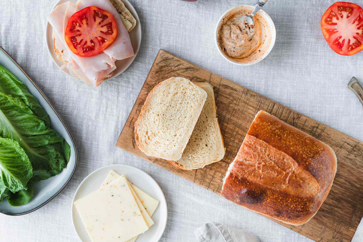 Sourdough Sandwich Loaf with Slices Cut on a Cutting Board, Surrounded by Sandwich Ingredients (a Small Plate with Cheese, a Platter with Lettuce, a Small Plate with a Slice of Bread Topped with Turkey Slices and Tomato Slice, a Small Bowl with a Sandwich Sauce, and a Sliced Tomato)