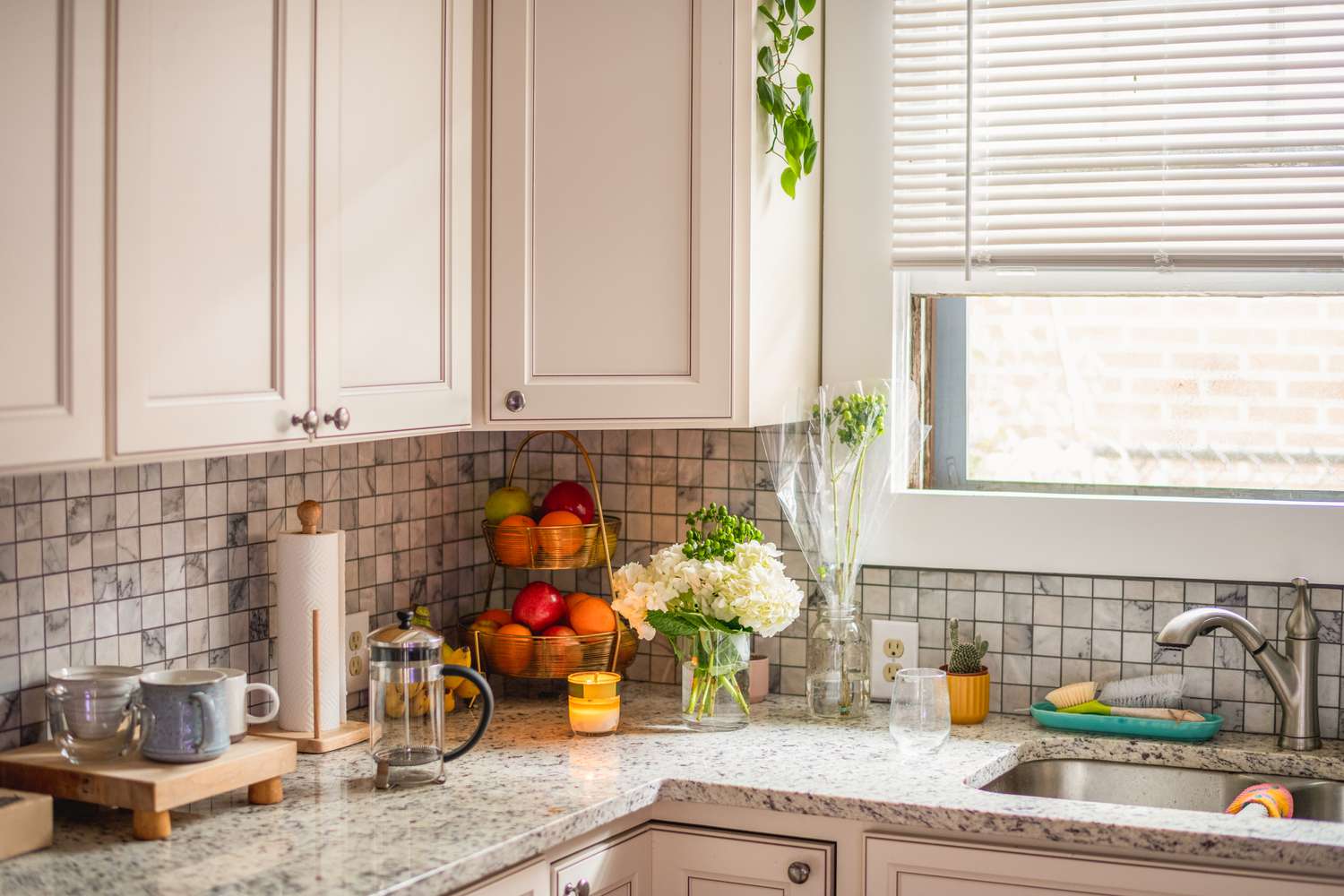 Photo of a clean kitchen, showing a kitchen sink with flowers, a basket of fruit, and a French press on the counter beside it