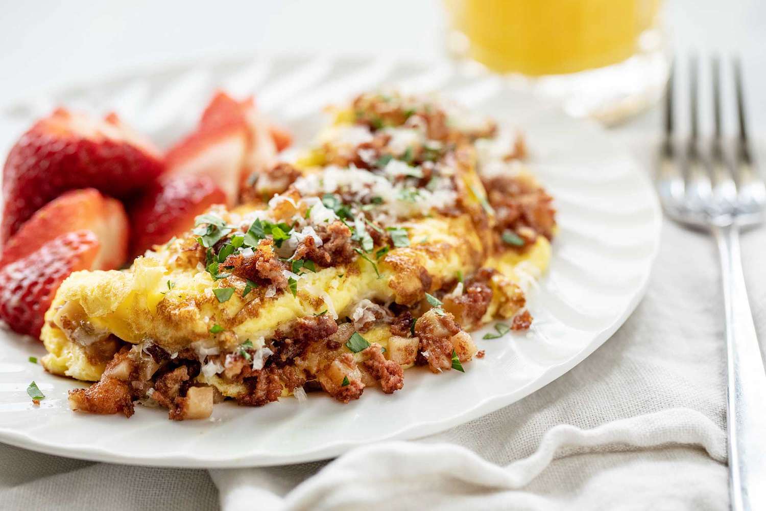 Horizontal image of the best corned beef omelet. The folded omelet is covered in browned corned beef. Grated cheese and parsley are on the top of the omelet. A white cotton napkin is partially under the plate. A fork rests on the napkin which is to the right of the plate. A partial view of a glass of orange juice is above the plate. Halved strawberries are to the left of the omelet on the plate.