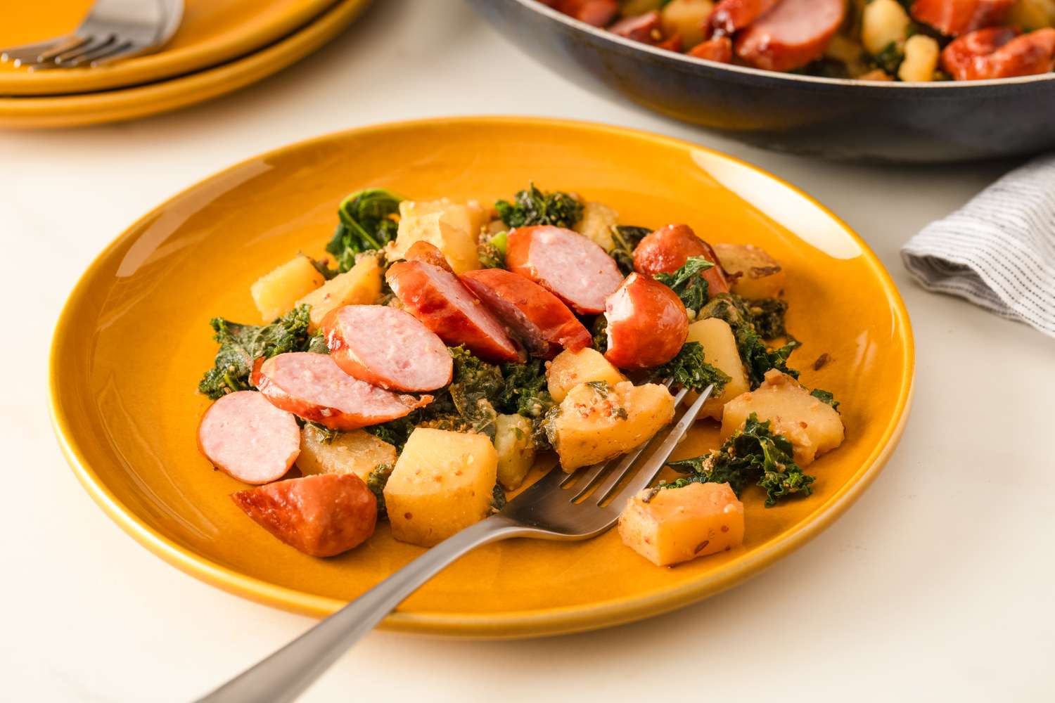 A plated dish of sausage slices potatoes and kale on a yellow plate with a fork