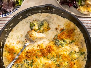Skillet chicken divan in a cast iron skillet (with some already served) at a table setting with a bowl of salad, a bowl of parmesan, a small bowl of chopped parsley, and a utensils