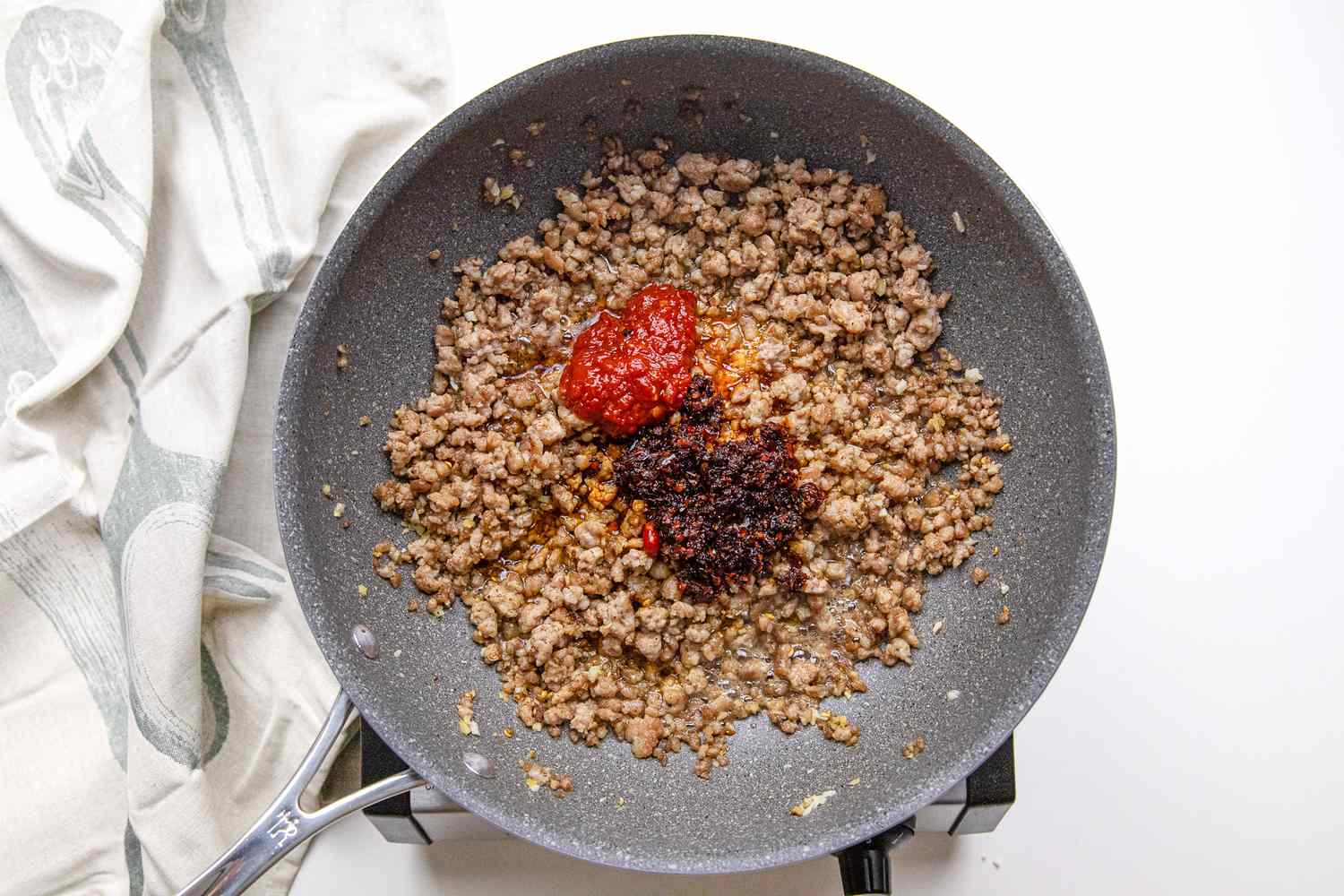 Chili Bean Paste and Chili Crisp Added to Ground Pork in a Pan on a Portable Burner Next to a White Kitchen Towel for Sichuan Mapo Tofu Recipe