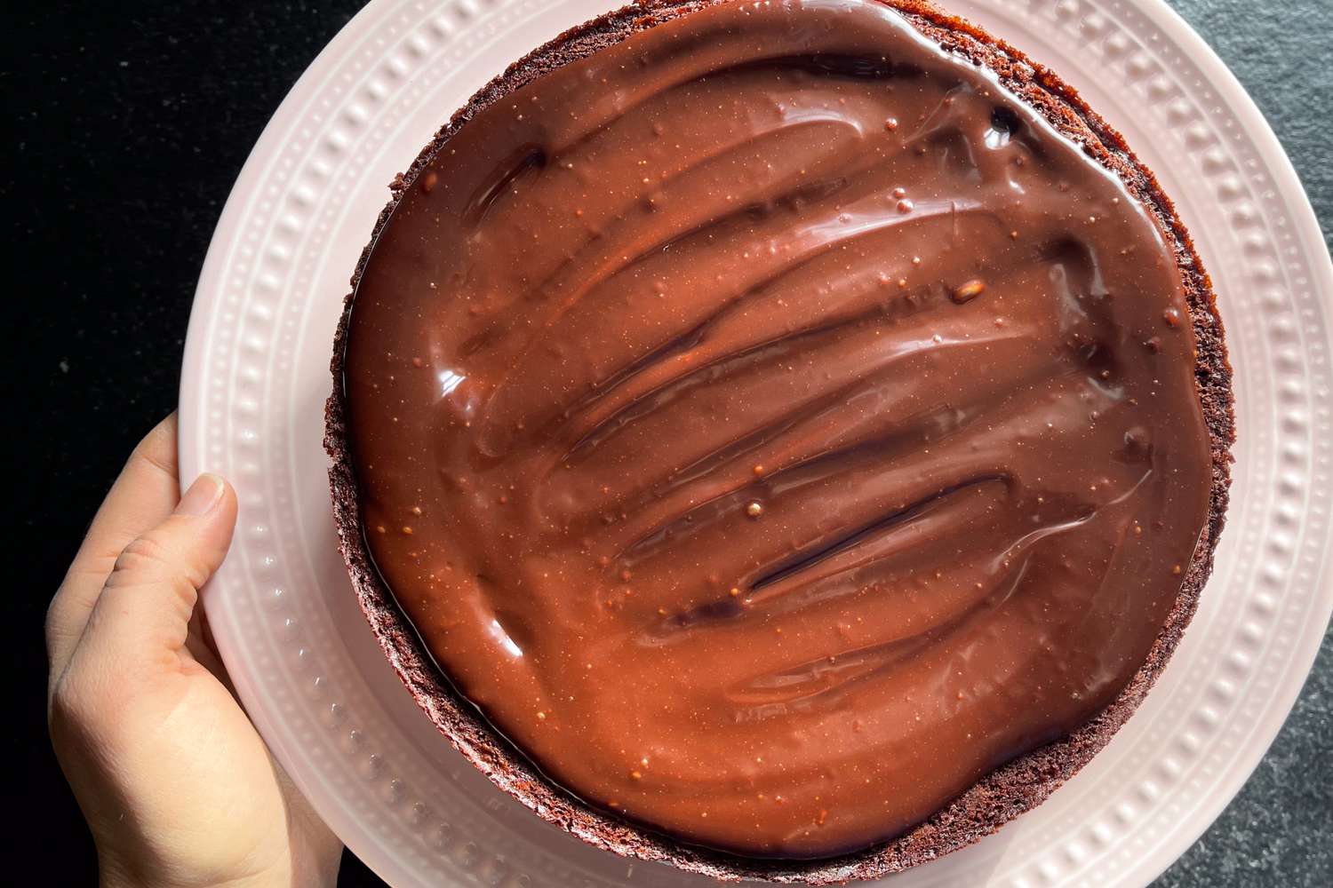 Hand holding a plate with a chocolate cake featuring a smooth thick chocolate glaze on top