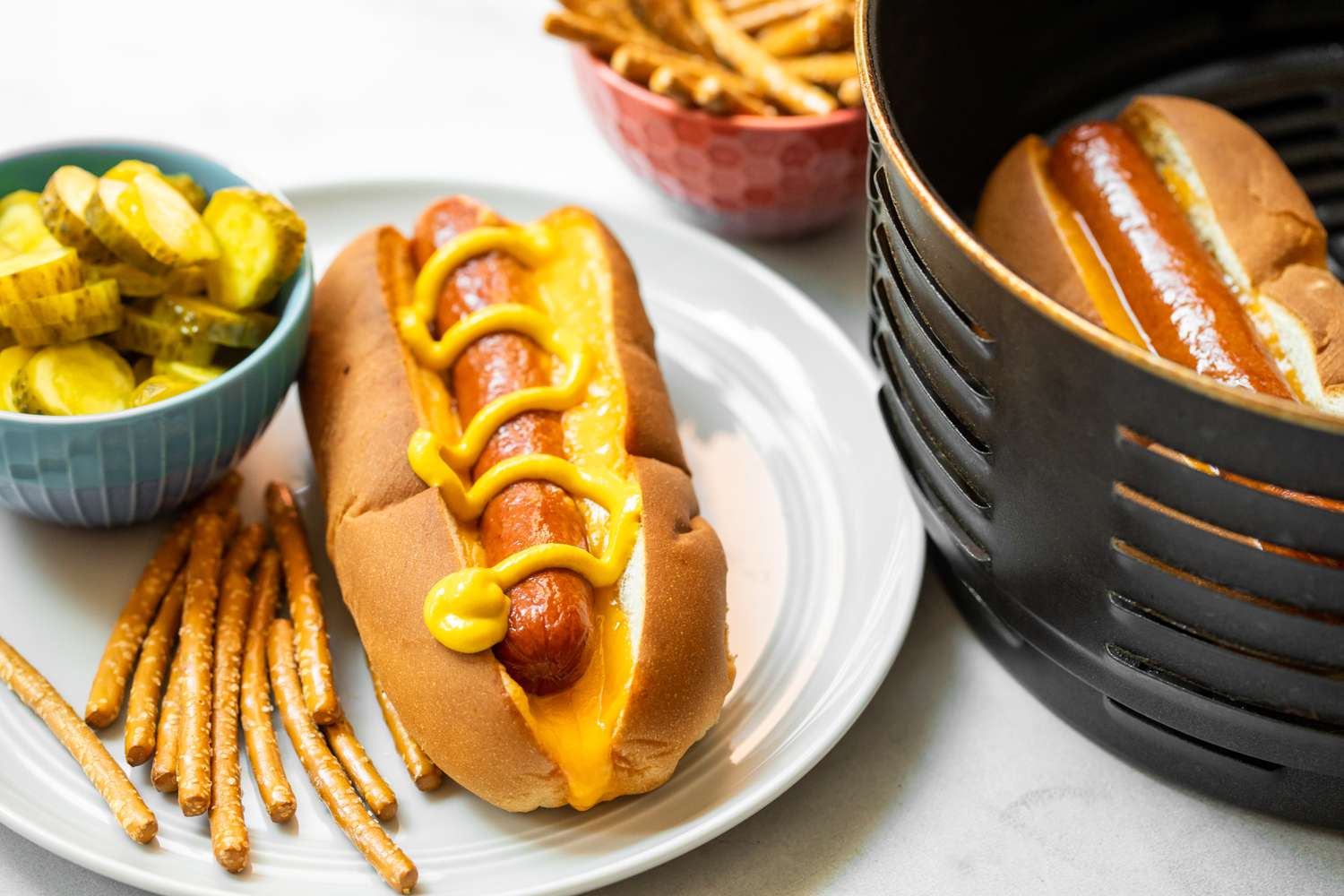A Plate of Air Fryer Hot Dog in a Bun Topped with Mustard and a Side of Pickles and Pretzels. Next to It Is the Air Fryer Basin with Another Hot Dog in a Bun and a Bowl of Pretzels.