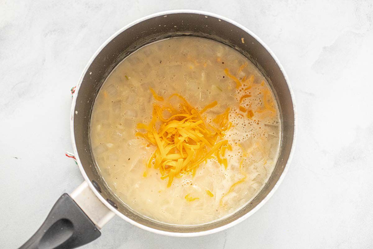 A saucepan with water, shredded cheddar cheese and shredded hash brown potatoes for Mormon Funeral Potatoes.
