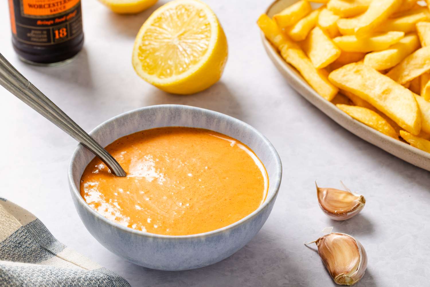 A bowl of sauce alongside french fries garlic cloves and a lemon half