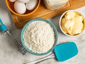 Baking ingredients and tools including flour eggs butter a whisk and a spatula on a countertop
