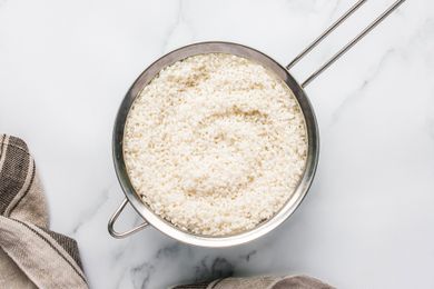 A bowl of rinsed rice sitting in a fine mesh strainer placed on a light surface with a towel nearby