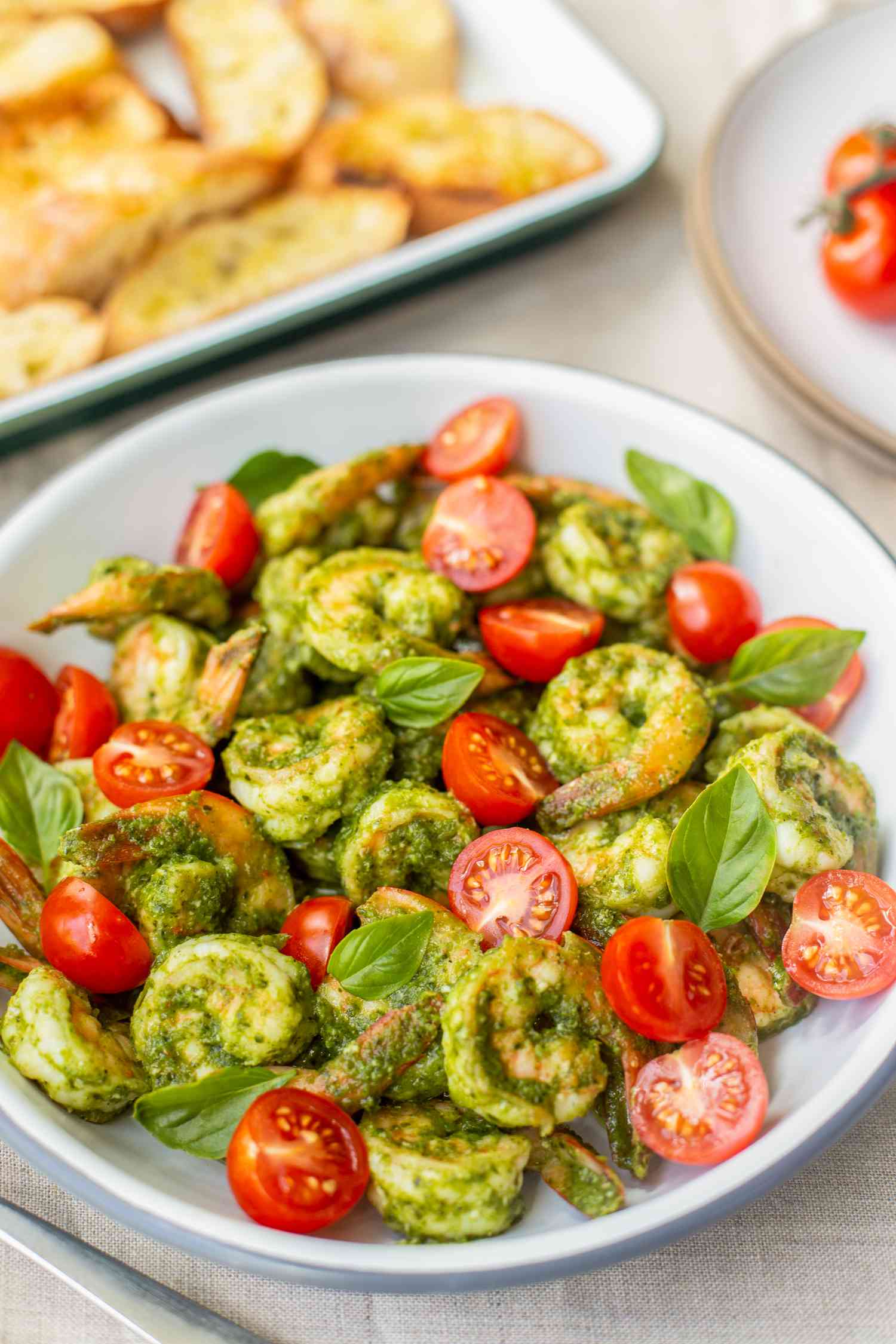 5-Ingredient Pesto Shrimp with Cherry Tomatoes in a Bowl, and in the Background, a Tray of Garlic Bread and Cherry Tomatoes