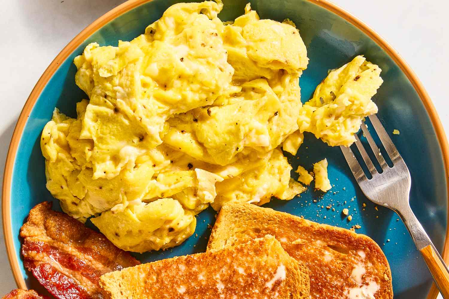Scrambled eggs served on a plate with toast and bacon a fork placed to the side